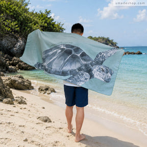 Umade 海洋微笑系列萬用快乾巾海龜款，站在沙灘上展示海龜圖案快乾巾，適合海邊玩水使用。SUP、游泳、衝浪、潛水、日光浴都適合，